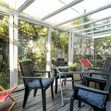 Ein gemütlicher Wintergarten mit schwarzen Stühlen und einem Tisch, umgeben von grünen Pflanzen. Ein roter Liegestuhl ergänzt die entspannte Atmosphäre.