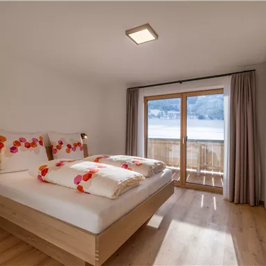 Ein helles Schlafzimmer mit einem großen Fenster und Zugang zum Balkon. Das Bett ist mit bunten Kissen dekoriert und der Boden ist aus Holz.