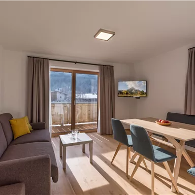 Ein modernes Wohnzimmer mit einem grauen Sofa und einem Holztisch. Große Fenster bieten Ausblick auf die Berge und sorgen für viel Tageslicht.