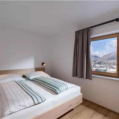 Ein gemütliches Schlafzimmer mit einem Doppelbett und modernen Bettdecken. Große Fenster bieten einen Blick auf die schneebedeckten Berge.