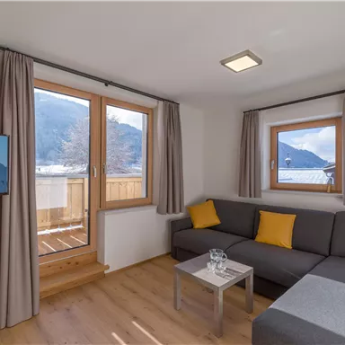 Ein modernes Wohnzimmer mit einem grauen Sofa und gelben Kissen. Große Fenster bieten einen Blick auf die Berge und viel Tageslicht.
