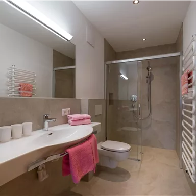Ein modernes Badezimmer mit einer Dusche und einem schlichten Waschbecken. Rosa Handtücher und eine helle, freundliche Atmosphäre prägen den Raum.