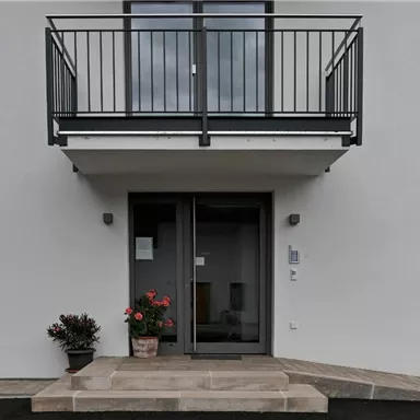 Ein modernes Gebäude mit einem Balkon und einer Eingangstür. Vor der Tür steht ein Blumentopf mit Blumen.