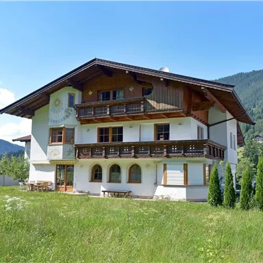Ein charmantes alpine Haus mit Balkonen und grünem Garten. Im Hintergrund sind sanfte Hügel und ein blauer Himmel zu sehen.