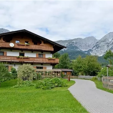 A traditional wooden house surrounded by green landscape and mountains. The sky is cloudy, and a path leads to the front door.