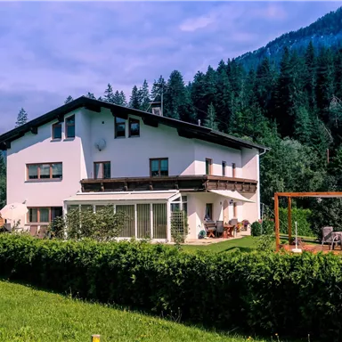 Ein modernes Haus umgeben von üppigem Grün und Bergen im Hintergrund. Die Terrasse lädt zum Entspannen ein.