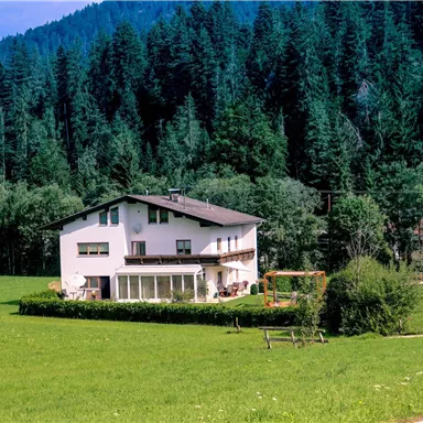 Ein gepflegtes Haus umgeben von grünen Wiesen und Bäumen. Im Hintergrund erheben sich die Berge unter einem klaren blauen Himmel.