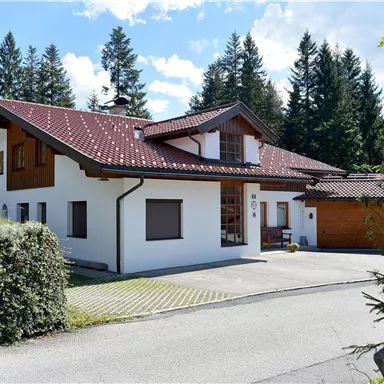 A modern house with a red roof and white facade, surrounded by trees. The entrance features an inviting driveway and parking space.