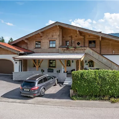 A large wooden house with a roof and a wide porch. In front of the house is a gray station wagon and there is a lot of greenery in the background.