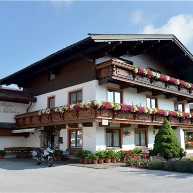 Ein traditionelles alpenländisches Haus mit einem schönen Balkon voller Blumen. Die Fassade ist hell und einladend, umgeben von grünem Garten.
