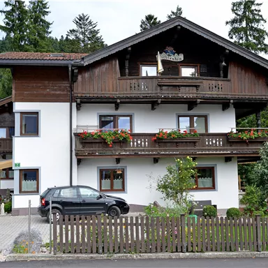 Ein zweigeschossiges Haus im alpinen Stil mit einem Balkon und blühenden Fenstern. Im Vordergrund steht ein Pkw und es gibt einen kleinen Garten mit einem Zaun.