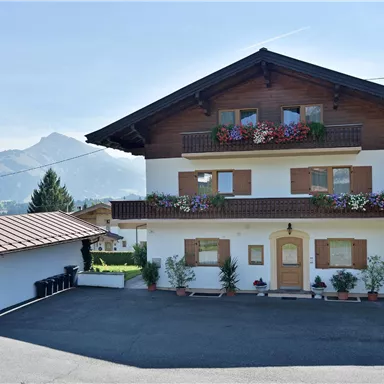 Ein zweistöckiges Chalet mit blühenden Balkonpflanzen und einer begrünten Umgebung. Im Hintergrund sind Berge zu sehen und der Himmel ist klar.