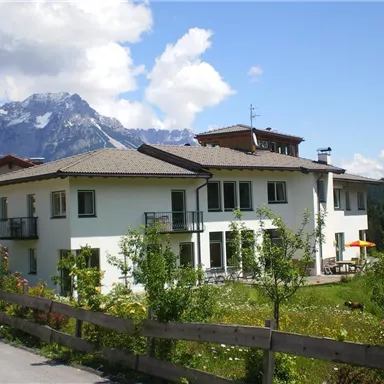 Ein modernes Haus mit Balkon, umgeben von grüner Landschaft und Blumen. Im Hintergrund sind die Berge sichtbar, unter einem blauen Himmel mit einigen Wolken.