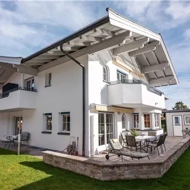A modern, two-story house with a spacious garden. The terrace is furnished with seating and offers a view of the surroundings.