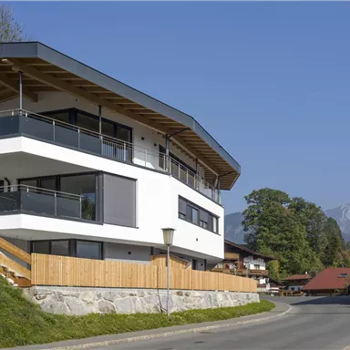 Ein modernes Haus mit einer großen Terrasse und Panoramafenstern. Im Hintergrund sind Berge und traditionelle Gebäude zu sehen.
