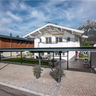 A modern house with a spacious carport and a well-maintained garden. In the background, impressive mountains can be seen and the sky is slightly cloudy.