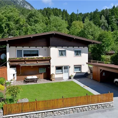 Ein schönes Haus mit einem gepflegten Garten und einem Carport. Umgeben von Bäumen und Bergen, präsentiert es eine ruhige und einladende Atmosphäre.
