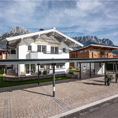 A modern house in the mountains, surrounded by green meadows and other chalets. The sky is blue and the mountain landscape is impressive in the background.