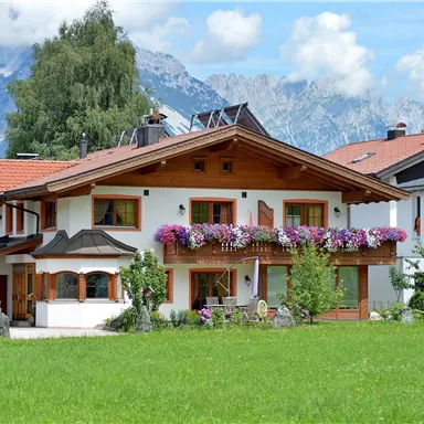 A charming house with a beautiful balcony, adorned with colorful flowers. In the background, impressive mountains and a blue sky can be seen.