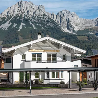A beautiful house with a modern design in front of impressive mountains. The surroundings are green and inviting.