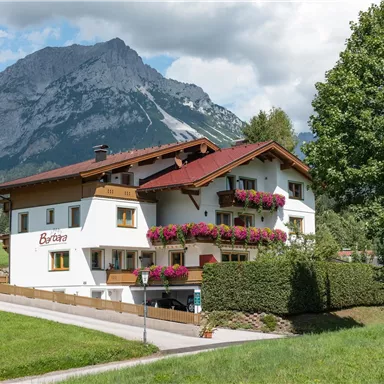 A picturesque house with flower-covered balconies, surrounded by green meadows. In the background, impressive mountains rise beneath a blue sky.