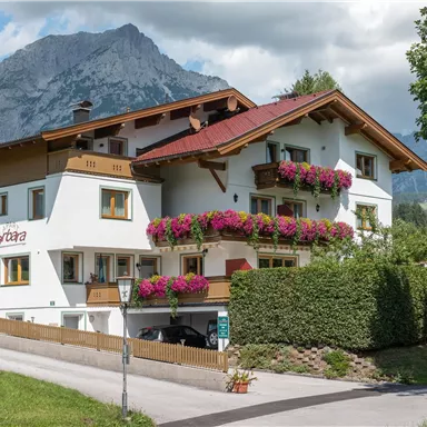 A charming house with colorful flowers on the balconies. In the background, majestic mountains and a blue sky can be seen.