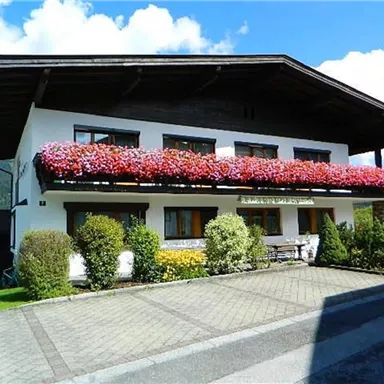 A pretty house with a blooming balcony and a well-kept garden. In the background, mountains and a blue sky can be seen.
