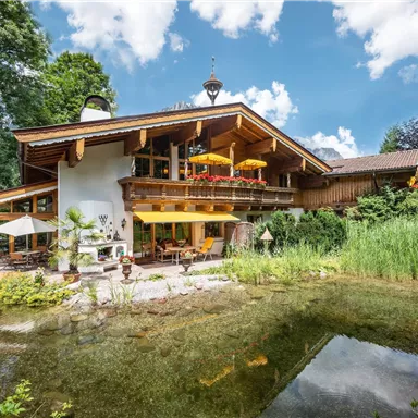 Ein charmantes Holzhaus mit einem schönen Garten und einem kleinen Teich. Im Hintergrund sind Bäume und ein strahlend blauer Himmel zu sehen.