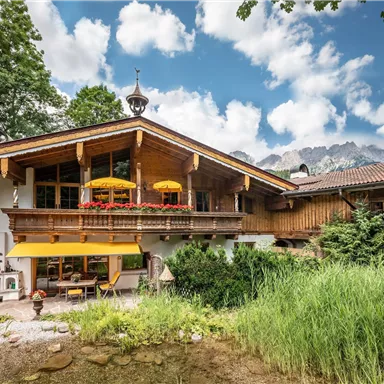 Ein charmantes Haus im alpinen Stil mit einem großen Balkon und bunten Sonnenschirmen. Umgeben von grünen Wiesen und einem klaren Himmel.