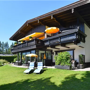 A beautiful house with a large garden and loungers. In the foreground, orange sun umbrellas can be seen.