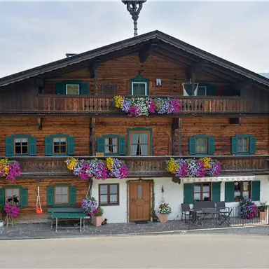 A traditional wooden house with colorful flowers on the balconies. In the background, gentle hills and a clear sky can be seen.