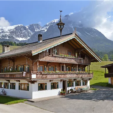 Ein traditionelles alpenländisches Haus umgeben von grünen Wiesen und hohen Bergen. Im Hintergrund sind schneebedeckte Gipfel sichtbar.
