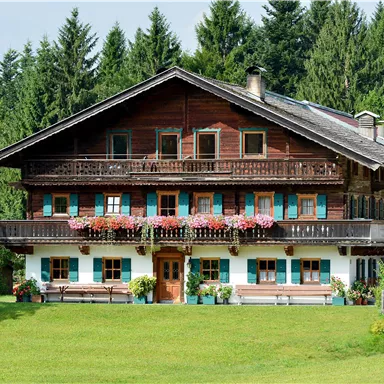 Ein traditionelles, rustikales Holzhaus mit Balkonen und bunten Blumen. Umgeben von einer grünen Wiese und bewaldeten Hängen.