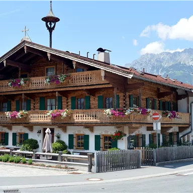 Ein traditionelles alpenländisches Haus mit mehreren Balkonen und bunten Blumen. Im Hintergrund sind Berge und ein blauer Himmel zu sehen.