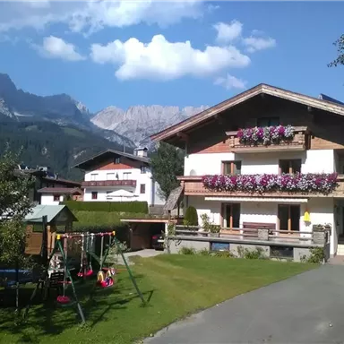 A charming house with blooming balcony flowers and a well-maintained garden. In the background, majestic mountains and a blue sky can be seen.