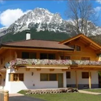 Ein schönes Haus mit einem blühenden Balkon und beeindruckenden Bergen im Hintergrund. Der Himmel ist klar und blau, was die malerische Landschaft unterstreicht.
