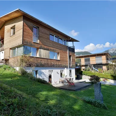 A modern wooden house in a green environment. In the background, mountains and a clear sky can be seen.