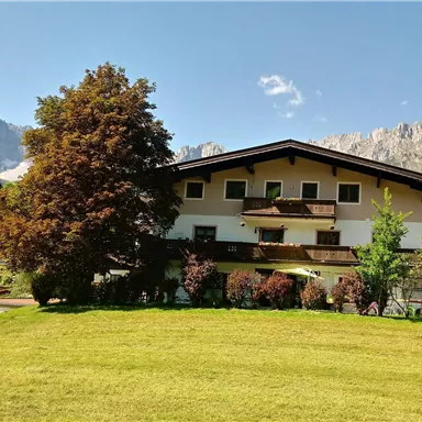 Ein malerisches Gebäude umgeben von einem grünen Garten und Bäumen. Im Hintergrund sind majestätische Berge zu sehen.