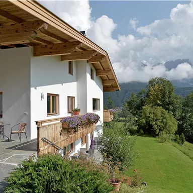 Ein modernes Haus mit einem großen Balkon und bunten Blumen. Im Hintergrund sind grüne Wiesen und Berge unter einem klaren Himmel zu sehen.