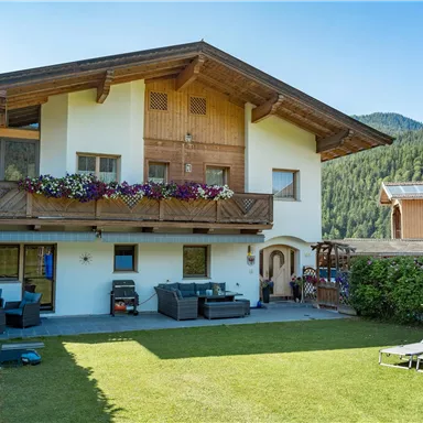 A cozy house with a garden in the mountains. The building has a balcony with flowers and is surrounded by green meadows.