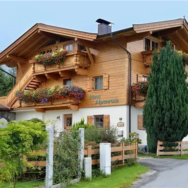A charming, wooden-clad house with flowered balconies and a well-maintained garden. The surroundings are adorned with green hills and a quiet path.