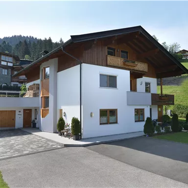 A modern house with wooden elements and a wide driveway. The surroundings are green with hills in the background.