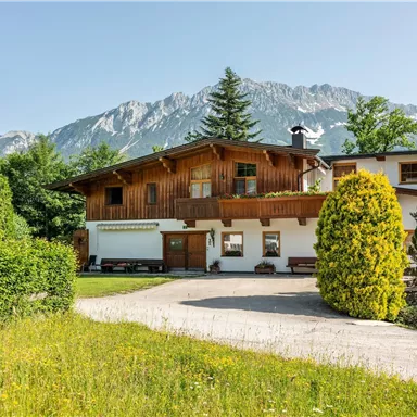 Ein charmantes Haus in einer malerischen Landschaft mit grünen Wiesen. Im Hintergrund sind hohe Berge und ein klarer Himmel zu sehen.