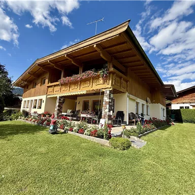 A beautiful house in Alpine style with a large garden and colorful flowers. The sky is clear and blue, which conveys a friendly impression.