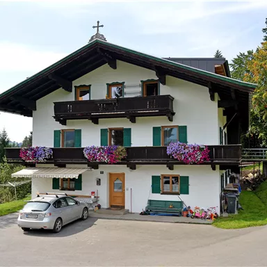A charming alpine house with blooming balconies. In front of the house stands a car, surrounded by a green landscape.