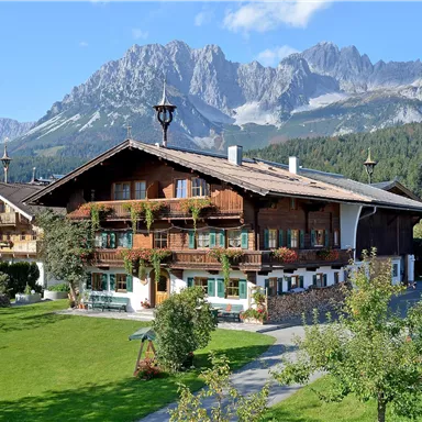 Ein traditionelles Holzhaus in einer grünen Landschaft. Im Hintergrund erheben sich majestätische Berge unter blauem Himmel.