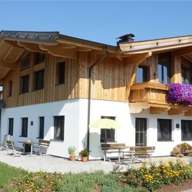 Ein modernes Holzhaus mit einem großen Garten und bunten Blumen. Im Hintergrund sind grüne Hügel und ein blauer Himmel zu sehen.
