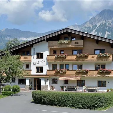 Ein charmantes Hotel mit blühenden Balkonen und einer schönen Berglandschaft im Hintergrund. Der Himmel ist klar und die Umgebung ist grün und einladend.