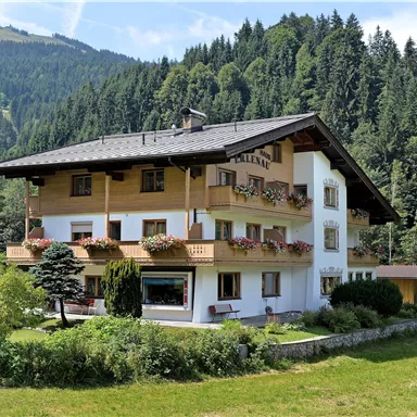 Ein charmantes Haus mit einem Holzbalcony inmitten von grünen Wiesen und Bäumen. Im Hintergrund sind Berge zu sehen, die eine malerische Atmosphäre schaffen.