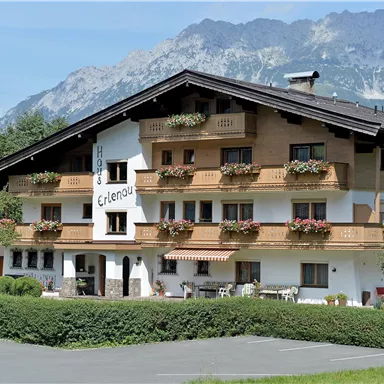 Ein charmantes Hotel mit Holzfassade und blühenden Balkonen. Im Hintergrund sind malerische Berge und ein klarer Himmel zu sehen.